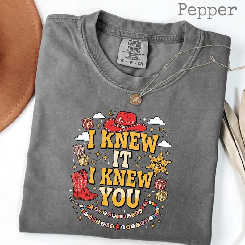 May include: A charcoal gray t-shirt with a graphic print featuring a red cowboy hat, red cowboy boots, alphabet blocks, and a sheriff star badge. The text reads I KNEW IT I KNEW YOU, EST. BESTIE 4EVA, and FOREVER FRIENDS on a beaded necklace design.