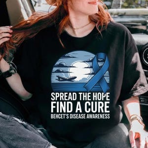 May include: Black t-shirt with a blue and white graphic. The graphic includes a blue ribbon, an airplane, and the text "SPREAD THE HOPE FIND A CURE BEHCET'S DISEASE AWARENESS". The design features a sky and ocean scene.