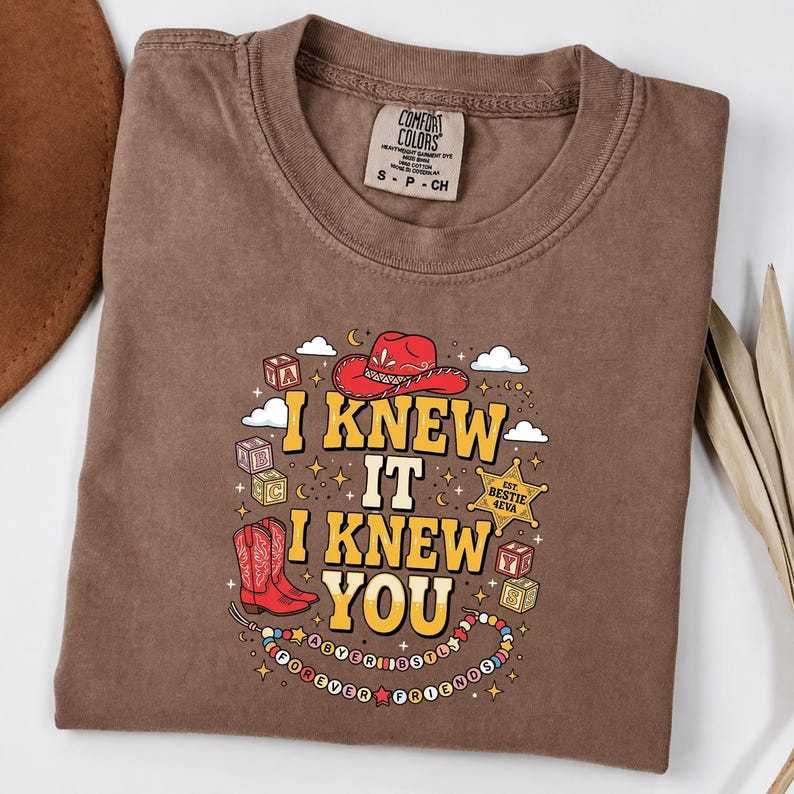 May include: A brown short-sleeve t-shirt featuring a graphic print with a red cowboy hat, red cowboy boots, alphabet blocks, and a gold sheriff star badge. The text reads I KNEW IT I KNEW YOU with a beaded necklace design and the words BESTIE 4EVA.