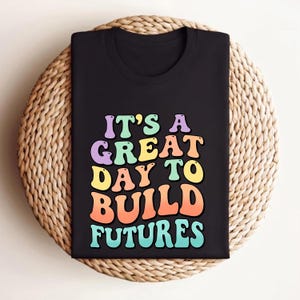 Puede incluir: Camiseta negra con el texto "IT'S A GREAT DAY TO BUILD FUTURES" en letras retro multicolores. La camiseta est&aacute; doblada y colocada sobre una superficie redonda tejida.