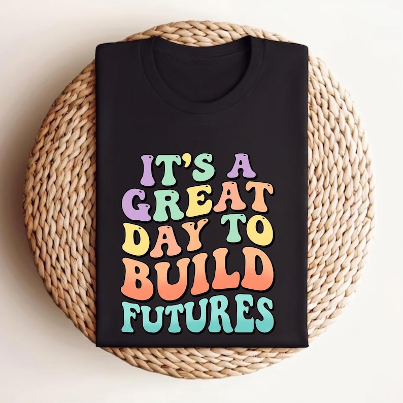 Puede incluir: Camiseta negra con el texto "IT'S A GREAT DAY TO BUILD FUTURES" en letras retro multicolores. La camiseta est&aacute; doblada y colocada sobre una superficie redonda tejida.