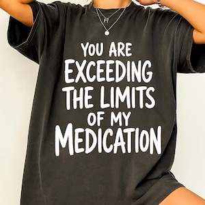 May include: A charcoal black short-sleeved t-shirt featuring bold white text printed on the front that reads: YOU ARE EXCEEDING THE LIMITS OF MY MEDICATION. The shirt has a crew neckline and a relaxed fit, showcasing a simple graphic design.