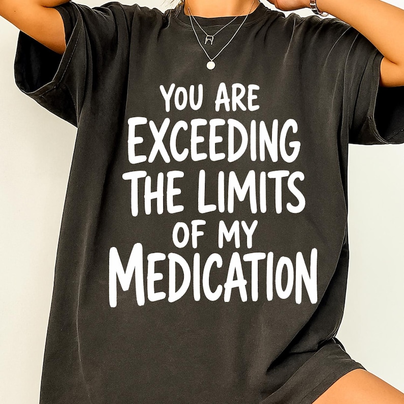 May include: A charcoal black short-sleeved t-shirt featuring bold white text printed on the front that reads: YOU ARE EXCEEDING THE LIMITS OF MY MEDICATION. The shirt has a crew neckline and a relaxed fit, showcasing a simple graphic design.