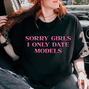 May include: Black t-shirt with the text "SORRY GIRLS I ONLY DATE MODELS" in pink. The shirt is a casual style, suitable for everyday wear. The text is centered on the front of the shirt.