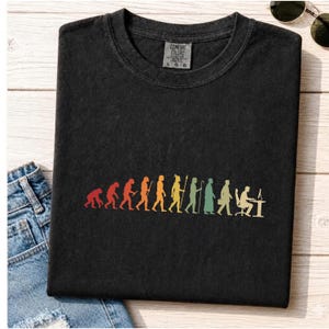 May include: A black t-shirt featuring a colourful graphic of human evolution, starting with a primate and progressing through various stages to a person sitting at a computer desk. The shirt has a crew neckline and a Comfort Colors brand label.