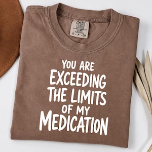 May include: A brown short-sleeved t-shirt with white text that reads "YOU ARE EXCEEDING THE LIMITS OF MY MEDICATION" printed on the front. The shirt features a crew neckline and a Comfort Colors brand label inside the collar area.