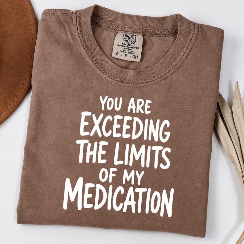 May include: A brown short-sleeved t-shirt with white text that reads "YOU ARE EXCEEDING THE LIMITS OF MY MEDICATION" printed on the front. The shirt features a crew neckline and a Comfort Colors brand label inside the collar area.