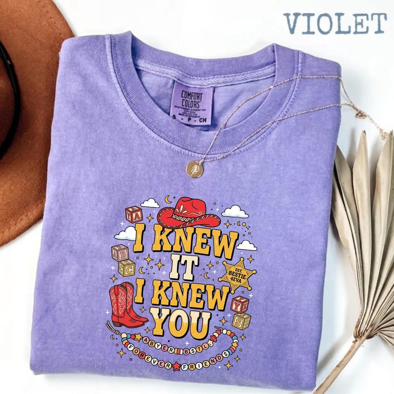May include: A violet short-sleeve t-shirt with a graphic print featuring a red cowboy hat, red cowboy boots, alphabet blocks, and a sheriff badge. The text reads I KNEW IT I KNEW YOU and FOREVER BESTIES. The shirt has a Comfort Colors brand label.