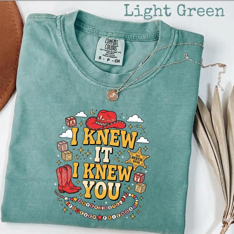 May include: A light green short-sleeve t-shirt featuring a graphic print with a red cowboy hat, red cowboy boots, alphabet blocks, and a sheriff badge. The text reads I KNEW IT I KNEW YOU, EST. BESTIE 4EVA, and FOREVER FRIENDS with colorful beads.