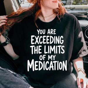 May include: A black short-sleeved t-shirt featuring bold white text that reads, "YOU ARE EXCEEDING THE LIMITS OF MY MEDICATION." The shirt has a crew neckline and a relaxed fit, with the graphic centered prominently on the front of the garment.