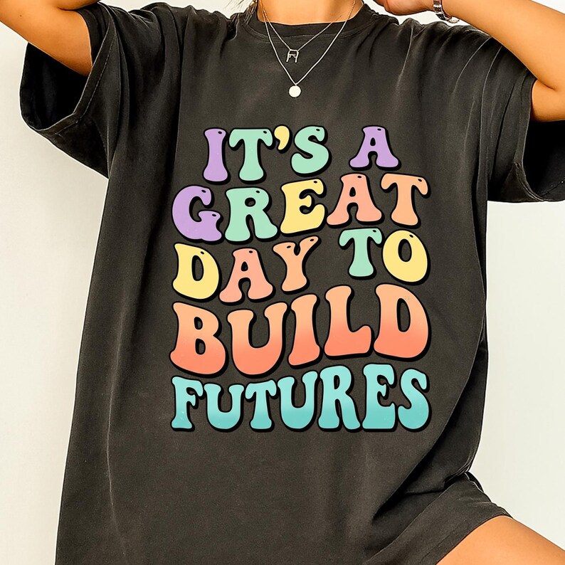 Puede incluir: Camiseta gris oscuro con el texto "IT'S A GREAT DAY TO BUILD FUTURES" en letras coloridas de estilo retro. La camiseta tiene un aspecto vintage y desgastado.