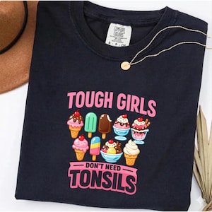 May include: A black t-shirt featuring the pink text TOUGH GIRLS DON'T NEED TONSILS and a graphic illustration of various ice cream cones, popsicles, and sundaes in shades of pink, brown, blue, and white, with a Comfort Colors brand label.
