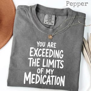 May include: A charcoal gray short-sleeve t-shirt with a crew neckline. The shirt features white text printed on the front that reads: YOU ARE EXCEEDING THE LIMITS OF MY MEDICATION. The garment has a Comfort Colors brand label inside the collar.