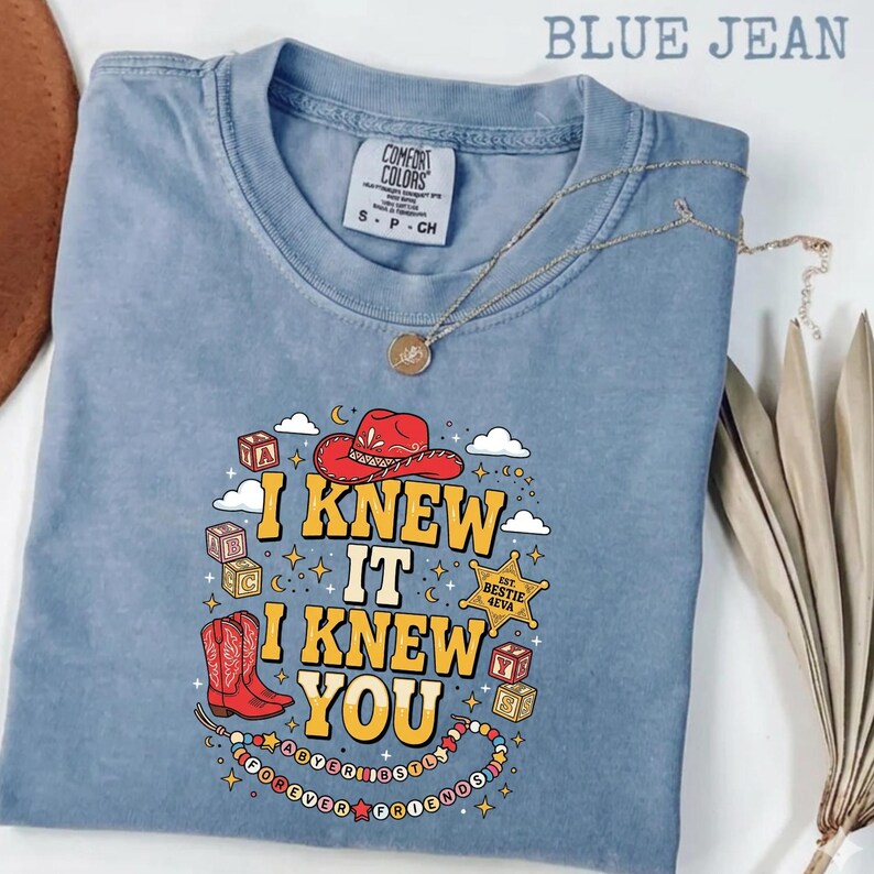 May include: A blue jean colored t-shirt featuring a graphic print with a red cowboy hat, red cowboy boots, alphabet blocks, and a sheriff star badge. The text reads I KNEW IT I KNEW YOU, EST. BESTIE 4EVA, and FOREVER FRIENDS. A thin gold necklace rests on the shirt.
