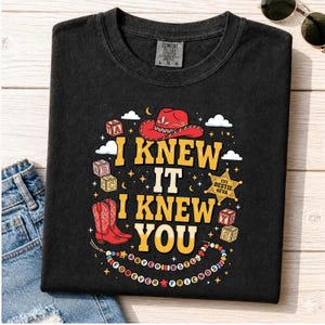 May include: A black t-shirt featuring a graphic print with the text "I KNEW IT I KNEW YOU". The design includes a red cowboy hat, red cowboy boots, alphabet blocks, a sheriff star badge labeled "EST. BESTIE 4EVA", and a beaded necklace with text.