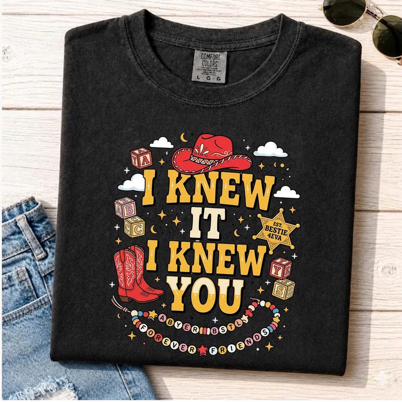 May include: A black t-shirt featuring a graphic print with the text "I KNEW IT I KNEW YOU". The design includes a red cowboy hat, red cowboy boots, alphabet blocks, a sheriff star badge labeled "EST. BESTIE 4EVA", and a beaded necklace with text.