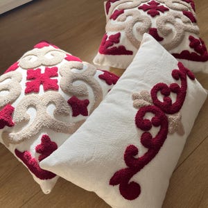 Peut inclure: Plusieurs coussins décoratifs avec un tissu blanc et des motifs brodés. Les coussins présentent des motifs rouges et beiges. Les coussins sont carrés et rectangulaires, disposés sur une surface en bois.