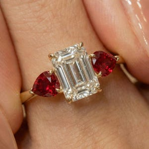 Puede incluir: Un anillo de oro con una gran gema clara rectangular flanqueada por dos gemas rojas en forma de pera. El engaste del anillo resalta el brillo de las piedras preciosas.