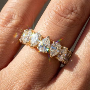 Könnte beinhalten: Ein goldener Ring mit einer Reihe von birnenförmigen Diamanten. Die Diamanten sind eng aneinander gesetzt und erzeugen ein durchgehendes Funkeln. Der Ring wird an einem Finger gezeigt, wobei sich das Licht in den Facetten der Diamanten spiegelt.