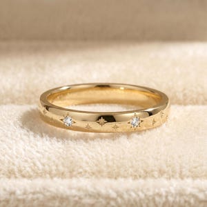 Könnte beinhalten: Ein goldfarbener Ring mit kleinen sternförmigen Gravuren und zwei größeren sternförmigen Fassungen mit klaren Edelsteinen. Der Ring wird auf einer weichen, cremefarbenen Oberfläche präsentiert. Die Ringbreite beträgt ca. 0,2 cm.