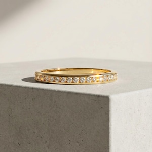 Könnte beinhalten: Ein goldener Bandring mit einer Reihe kleiner, klarer Edelsteine, die rundherum eingesetzt sind. Der Ring wird auf einer grauen Oberfläche präsentiert, wobei das weiche Licht seine polierte Oberfläche hervorhebt. Der Ring ist ein Schmuckstück.