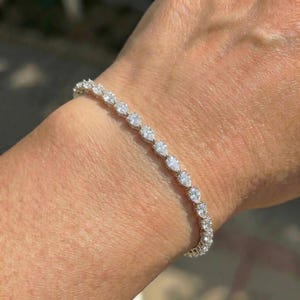 Pode incluir: Uma pulseira delicada com uma série de pedras preciosas claras em forma de pêra. As pedras são colocadas próximas umas das outras, criando uma linha contínua de brilho. A pulseira é usada no pulso, mostrando seu design elegante.