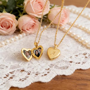 May include: Gold-colored heart-shaped locket necklaces. One locket is open, revealing two small photos. The other locket has the letter "A" engraved on the front. The necklaces are displayed on a white lace surface with pink roses and a string of pearls.