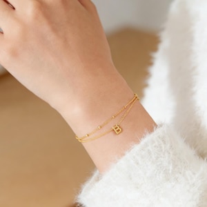 Könnte beinhalten: Ein goldfarbenes, zweilagiges Armband mit einem kleinen Buchstaben "B" Anhänger. Das Armband hat zarte Perlenakzente entlang der oberen Schicht und wird am Handgelenk getragen. Der Hintergrund ist verschwommen, mit einem weißen Pullover sichtbar.