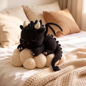 Peut inclure: Un jouet en forme de dragon noir crocheté, avec des cornes, des ailes et une base en forme de nuage crème. Le dragon est allongé, les yeux fermés. Le jouet est sur un lit avec des oreillers et une couverture.