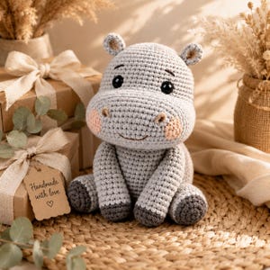 Peut inclure: Un hippopotame en peluche crocheté gris clair avec des pieds gris foncé, de petites oreilles arrondies et des joues rose pâle. L'hippopotame a des yeux de sécurité noirs et une bouche cousue. Une étiquette cadeau en papier brun indique Handmade with love.