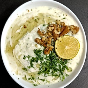 May include: A white bowl filled with a creamy white dip or soup, topped with a cluster of brown walnut halves, a bright yellow lime slice, and a generous sprinkling of fresh green dill, with a drizzle of clear oil visible on the surface.