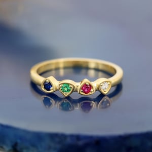 Op de afbeelding: Een gouden ring met vier kleine, ronde edelstenen die in een rij zijn gezet. De stenen bestaan uit een diepblauwe saffier, een levendige groene smaragd, een helderrode robijn en een heldere witte diamant, gezet in een gepolijste gouden band.