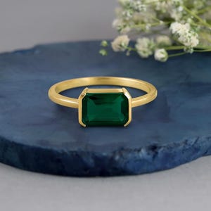 Peut inclure: Une bague en or ornée d'une pierre précieuse verte de taille émeraude rectangulaire, fixée dans un serti clos lisse. L'anneau est fin et poli, présentant un design épuré qui met en valeur la couleur vive de la pierre centrale.