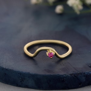 Puede incluir: Un anillo de oro con una banda lisa y curva que presenta una única piedra preciosa redonda de color rosa vibrante en un engaste de garras. La banda tiene una suave forma ondulada que eleva la piedra, creando una pieza de joyería sencilla.
