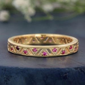May include: A gold band ring featuring a repeating geometric triangle pattern along the outer surface. Small, round, vibrant pink gemstones are set within alternating triangles, creating a continuous decorative motif around the entire circumference.
