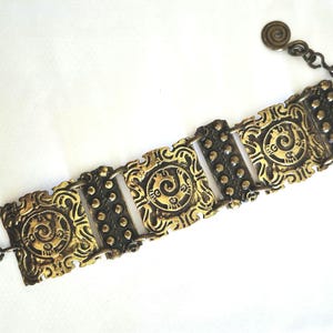 Peut inclure: Un bracelet en métal doré composé de trois panneaux carrés à motifs de spirales en relief alternant avec deux panneaux rectangulaires ornés de rangées de petits points en relief. Il comprend un fermoir circulaire et une breloque spirale.
