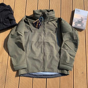 Könnte beinhalten: Olivgrüne Gore-Tex-Jacke mit durchgehendem Reißverschluss, hohem Kragen und mehreren Taschen. Die Jacke hat Aufnäher auf den Schultern und ein Etikett mit der Aufschrift "Gore-Tex". Links ist ein schwarzes Kleidungsstück zu sehen, rechts ein Buch mit dem Titel "FULL STOP".