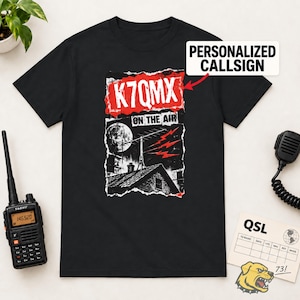Può includere: Una t-shirt nera a maniche corte con una stampa grafica di un tetto di casa con antenna, una luna e fulmini rossi. Il testo recita K7QMX ON THE AIR. Una freccia rossa punta al nominativo con il testo PERSONALIZED CALLSIGN.