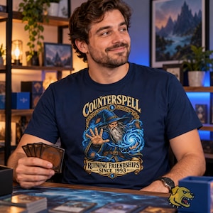 Könnte beinhalten: Ein marineblaues T-Shirt mit einer Grafik eines Zauberers in blauem Gewand und Hut, der einen Zauberspruch wirkt. Der Text "Counterspell" steht oben und "Ruining Friendships Since 1993" unten, umrahmt von einer verzierten gold-blauen Bordüre.