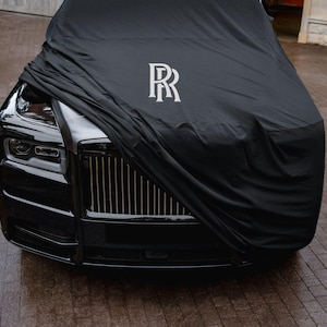 Peut inclure: Une voiture noire recouverte d'une housse noire. La housse porte un logo blanc. La voiture est garée sur une surface en briques.
