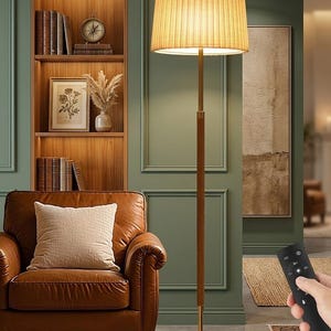 May include: A tall floor lamp with a pleated lampshade, wooden pole, and a brass base. The lamp is in a room with a leather armchair, a bookshelf, and a painting. A remote control is in the foreground.