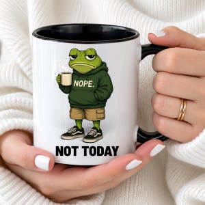 May include: A white ceramic mug with a black interior and handle, featuring a graphic of a green frog wearing a dark green hoodie with the text NOPE. The frog holds a small mug, and the text NOT TODAY is printed in black below the illustration.