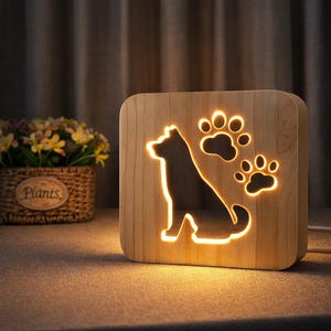 Könnte beinhalten: Ein Nachtlicht aus Holz mit einer Hunde-Silhouette und Pfotenabdrücken. Das Licht leuchtet von innen durch den Holzrahmen und erzeugt eine warme Atmosphäre. Das Licht ist quadratisch.