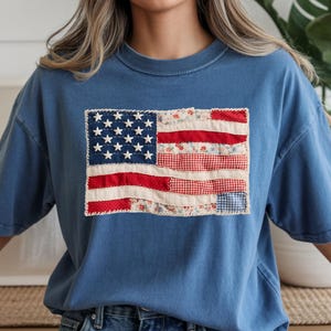 May include: A blue t-shirt featuring a patchwork American flag design. The flag incorporates red, white, and blue fabrics with floral, gingham, and solid patterns. The shirt has short sleeves and a crew neck.
