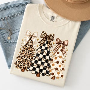 Może przedstawiać: Kremowy t-shirt z grafiką przedstawiającą trzy stylizowane choinki. Pierwsze drzewko ma wzór w panterkę z kremową kokardą, drugie czarno-białą szachownicę z kokardą w panterkę, a trzecie wzór w kropki z brązową kokardą w kratę.