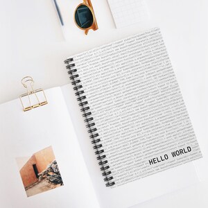 Minimalist white spiral notebook with ‘Hello World’ text and code pattern cover, developer-themed stationery on white desk.
