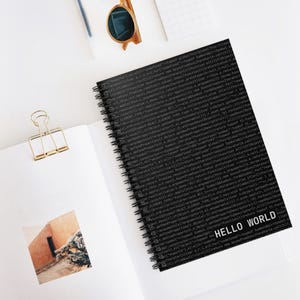 Minimalist black spiral notebook with ‘Hello World’ text and code pattern cover, developer-themed stationery on white desk.