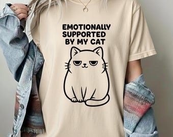 May include: Beige t-shirt with the text "Emotionally Supported By My Cat" above a black cartoon cat illustration. The cat has a grumpy expression. The shirt has short sleeves and a crew neck.