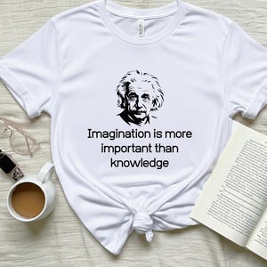 Pode incluir: T-shirt branca com uma ilustração a preto e branco de Albert Einstein e a citação "A imaginação é mais importante que o conhecimento". A t-shirt é feita de tecido macio, ideal para uso diário.
