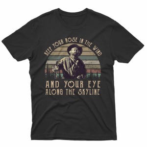 May include: Black t-shirt featuring a vintage-style graphic of a man in a hat holding a rifle, set against a sunset backdrop. The text reads "KEEP YOUR NOSE IN THE WIND AND YOUR EYE ALONG THE SKYLINE."